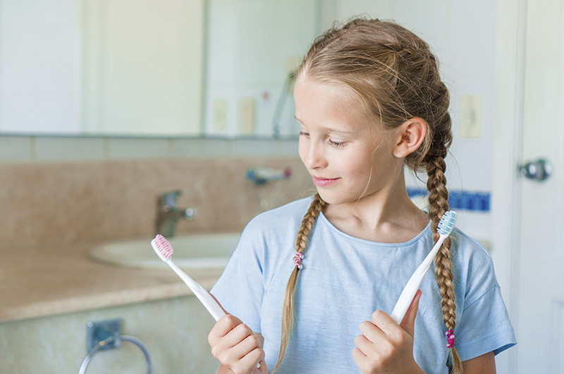 A imagem mostra uma menina branca, de cabelos loiros e com duas tranças. Ela está em um banheiro e veste uma camiseta azul. Em cada mão segura uma escova de dentes e olha para a que está à sua direita.