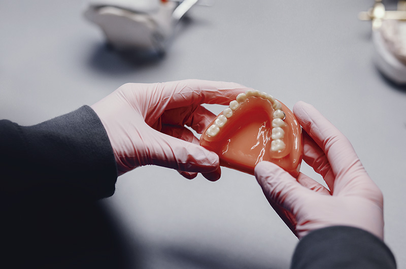 no centro da imagem, mãos usando luvas de látex seguram uma prótese dentária total superior. O fundo da imagem é cinza e tem alguns elementos desfocados.