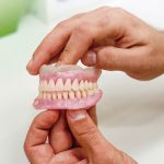 Na imagem uma pessoa segura em suas mãos uma prótese de arcada dentária. É possível ver todos os dentes do lado esquerdo do objeto.
